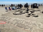 A sand sculpture of a large sea creature with tentacles stretching out, with a message reading "HAPPY BIRTHDAY ERIC AND TEDDY" in the foreground. Beach chairs are set up to the left of the competition plot, with spectators in the background.