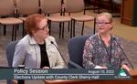 Two women sit at a desk at a televised public meeting.