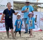 Three children hold up ribbons wearing medals. They're all smiling big before the event stage, and some are wearing blue event t-shirts. They stand before the event stage in the sand.