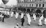 People wearing white gowns and hoods march openly in a parade through a downtown street in a black and white photo.