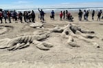 A sand sculpture of a big lobster, octopus and sea turtle, all facing each other in the center. Written in the sand in partial view is "Rock, Paper, Scissors." Spectators watch and take pictures in the background.