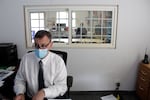 A person in a white dress shirt with a tie sits in a chair in an office with a mask on his face.