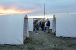 A small crowd mills between rows of huts that are covered by white tarps.