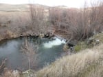 The river water foams at the base of the dam, which is surrounded by sagebrush and wild grasses on a cloudy day.