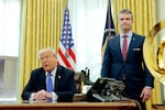 In this photo, President Trump is seated at his desk in the Oval Office, and standing beside the desk is Defense Secretary Pete Hegseth. Both men are wearing suits and ties.