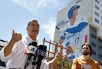 Xavier Becerra is pictured gesturing with news microphones in front of him. In the background, blue sky is visible as well as a huge mural of LA Dodger baseball star Shohei Ohtani.