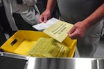In this file photo from 2024, Pennsylvania poll workers demonstrate how ballots are are received, processed, scanned, and securely stored. This image shows a box of yellow ballots as a poll worker holds one with a ballot peeking out. 