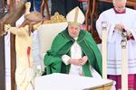 Pope Francis, dressed in green and white, sits next to a crucifix and looks to the side.