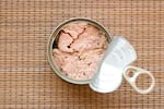 This photo, taken from above, shows a can of tuna. The lid, which has a pull tab, is peeled back, revealing the tuna in the can. The can sits on a rattan-like mat.