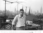 Beat poet Allen Ginsberg, photographed in black-and-white on a rooftop by William S. Burroughs in 1953. The young Ginsberg wears thick framed eyeglasses, a white button-up shirt with the sleeves rolled up, slacks and a serious expression. Behind him in view are rooftops and a distant cityscape.