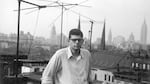 Beat poet Allen Ginsberg, photographed in black-and-white on a rooftop by William S. Burroughs in 1953. The young Ginsberg wears thick framed eyeglasses, a white button-up shirt with the sleeves rolled up, slacks and a serious expression. Behind him in view are rooftops and a distant cityscape.