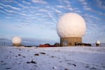 Three buildings with white globe shapes on top of them at Pituffik Space Base are shown in a frozen landscape with snow on the ground and underneath a bright blue sky striped with clouds in northern Greenland.