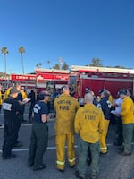 A group of people in shirts that say "Gresham Fire" and "Portland Fire" on their backs stand in a Southern California parking lot by four red fire engines. The sky is blue and palm trees are seen in the background.