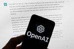 OpenAI's logo is on the screen of a mobile phone that's being held in front of a computer screen displaying output from ChatGPT.