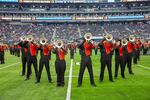 Band members in orange and black uniforms stand on a football field. Their faces are blocked by shiny silver instruments.