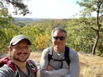 Rick Colby, 64, right, wears sunglasses . His 32-year-old son Ashton, left, wears a baseball cap. They both wear backpacks and are photographed with a backdrop of trees and mountains.