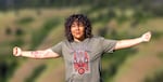 A Native American woman with curly, shoulder-length dark brown hair wears a gray t-shirt with her arms stretched out and her fists balled. The red handprint tattoo on her right arm, a symbol of Murdered and Missing Indigenous Women, is joined by doves representing members of her family who have died.