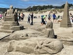 This sand sculpture features a cat seated on a throne and a pointed tower with Egyptian hieroglyphs at the base, and in the foreground, an open coffin with a mummy inside sitting up. Spectators are looking on and taking photos in the background.