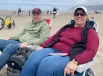 A woman wearing sunglasses, warm vest and a white baseball cap, and a man wearing a sweatshirt and gray baseball cap, smile seated in beach chairs on the beach.