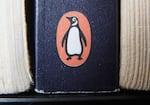 The Penguin logo is visible on the spine of a book. The U.S. Department of Justice is suing Penguin Random House and Simon & Schuster to block the companies from completing a merger valued at $2.175 billion.