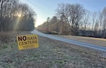 A yellow sign saying "NO DATA CENTERS for Walnut Cove... the Dan River & Stokes County" is staked into the ground by the side of a rural road with dried grass and leafless trees on each side.
