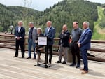 A man speaks at a podium, surrounded by five other men and one woman, on an outdoor patio overlooking a grassy, forested ski slope in summer.