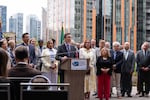 Washington Gov. Bob Ferguson speaks from behind a podium at an open-air press conference, surrounded by more than a dozen people with downtown Seattle buildings providing a backdrop.