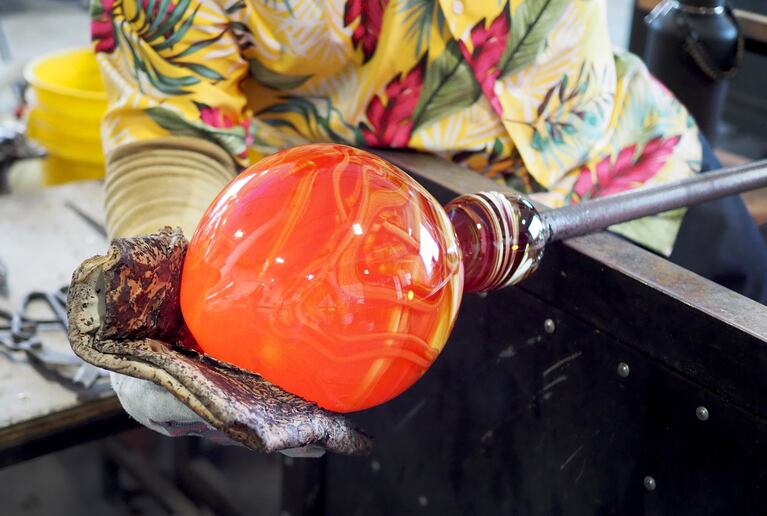 A man holds an orange ball of hot glass in an oven-mitted hand.