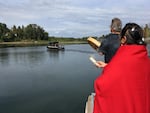 Two people in the foreground watch a boat as it travels across a river.