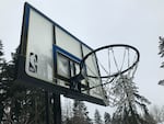 The glass backboard of a basketball hoop has ice accumulation on a cloudy day.