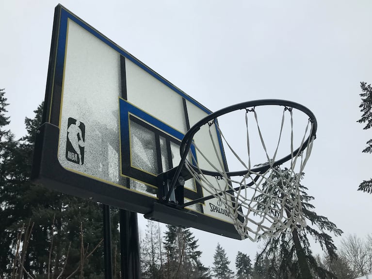 The glass backboard of a basketball hoop has ice accumulation on a cloudy day.