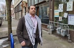 Man stands in street - spots coat over pink collared shirt and white and black scarf draped over shoulders