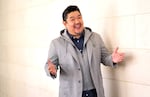Voice actor Eric Bauza, wearing a gray suit jacket and holding out his arms, poses for a portrait next to a cream colored wall.