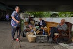 An elderly woman holding leafy vegetables and a cane walks past two street vendors selling farm produce and traditional goods under an overpass in Chongqing, China.