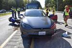 This photo shows the front of a dark gray Tesla sedan on a road after a fatal collision with a motorcycle. On the left side of the photo, the motorcycle lies on its side on the road, next to the Tesla. First responders wearing yellow vests assess the scene.