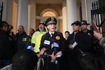 A police officer in uniform speaks to a press conferene with other law enforcement including FBI outdoors in fornt of a building with pillars.