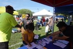 A man in a bright yellow shirts shakes the hand of a grinning man in sunglasses in front of a small booth. At the booth, a committee member organizes paperwork while a woman fills out voter registration forms.