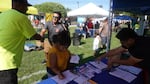 A man in a bright yellow shirts shakes the hand of a grinning man in sunglasses in front of a small booth. At the booth, a committee member organizes paperwork while a woman fills out voter registration forms.
