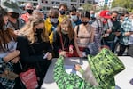A person wearing a top had and suit jacket covered in images of cannabis plant leaves hands out joints to a crowd of people wearing  masks.