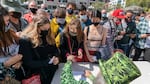 A person wearing a top had and suit jacket covered in images of cannabis plant leaves hands out joints to a crowd of people wearing  masks.