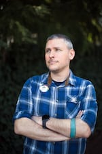 Joshua Morris, photographed from about the waist up, stands with his arms crossed over his waist. He's wearing a blue plaid shirt with sleeves rolled up just past the elbows. Pinned to his shirt is a button that says Birds Connect Seattle and that bears the nonprofit's logo.