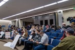 People sit in auditorium style seats. Most ear masks, but a woman in the front row has her face covering lowered below her mouth. Many people carry signs. One reads: "heart not lockdown." Another sign reads "consent through coersion will backfire no to any mandate"