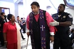 Rev. Dr. William Barber is wearing a purple stole embroidered with a colorful cross, has a red armband tied around his left arm, and leans on a cane with his right arm while he is being arrested.