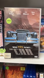 A front-facing DVD box is seen on a white shelf among other movies. It features the shiny grill of a classic car, with eerie red-tinted scenery in the background. Above the movie title, The Car's tagline reads, "WHAT EVIL DRIVES...."