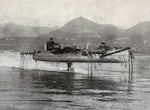 A black and white photo of an elevated canoe-like boat over a lake with people in it.