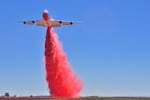 An airplay deploys a plume of reddish-pink fire suppressant from its tail.