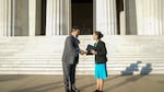 Sunlight colors the Lincoln Memorial steps light yellow. Charles Sams stands on the left in a dark gray suit and blue shirt, a beaded medallion hanging from a necklace on his chest and an eagle feather in his hair. Deb Haaland stands on the right in a turquoise dress, black blazer and turquoise jewelry. Each is smiling as they shake hands, Haaland holding a black book in her left hand. The Abraham Lincoln statue is covered in shadow and visible in the background.