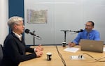 A woman and a man sit across a conference table and talk into microphones.