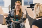 In an office setting, an Asian American woman smiles at a colleague. The woman has long brown hair. She sits in a wheelchair. 