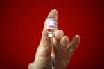 A hand wearing a medical glove holds a vial labeled "AstraZeneca," while a syringe enters into the vial.